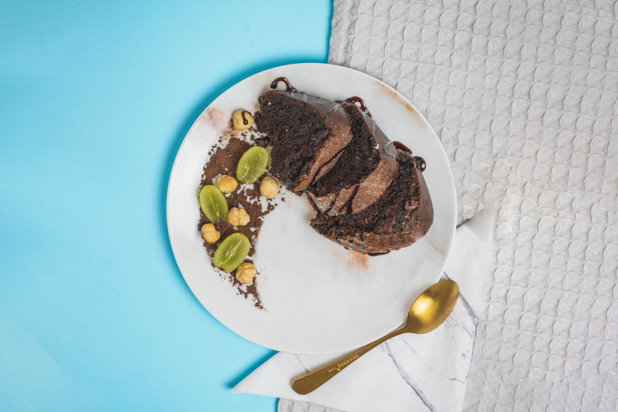 Kousky kakaové bábovky přelité čokoládou na bílém talíři, dozdobené práškovým kakaem, ořechy a hroznovým vínem